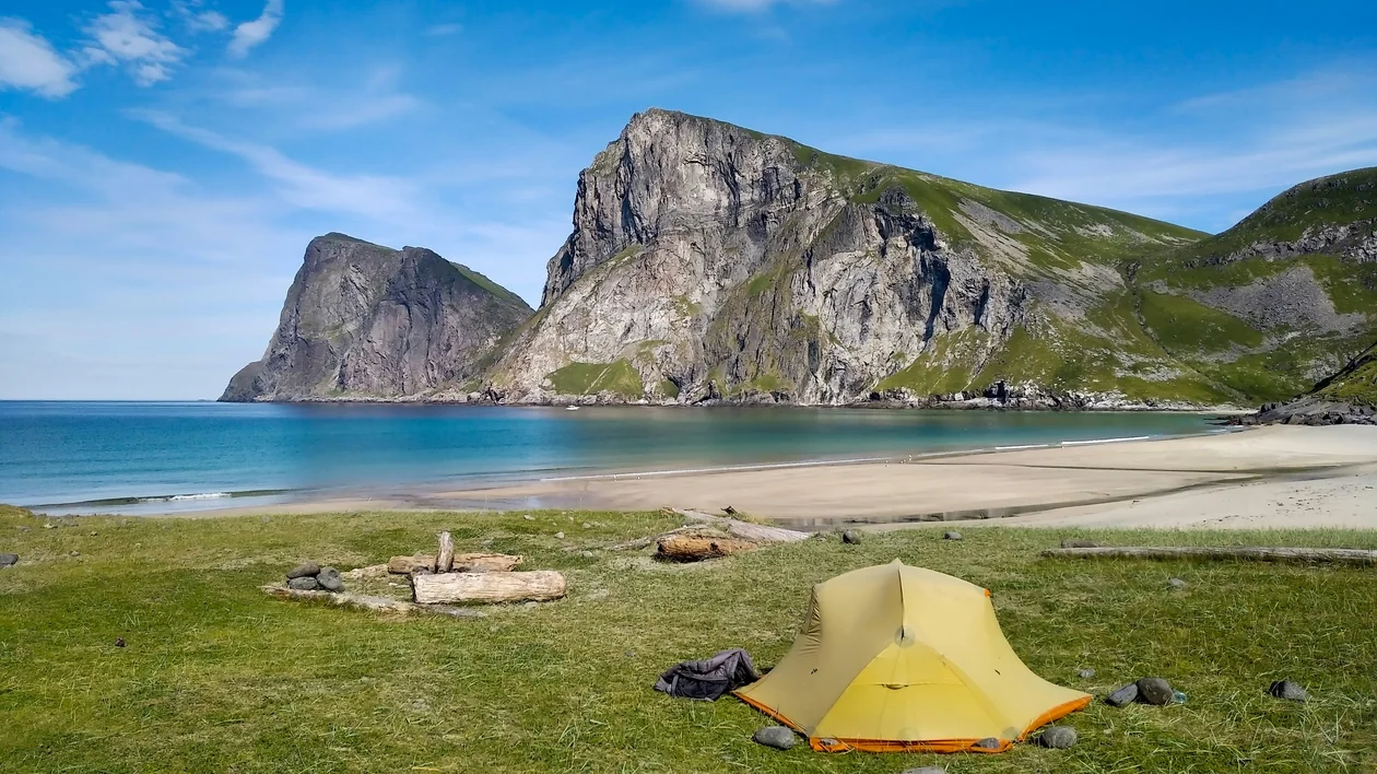 Yellow tent on grassy shore with mountains and blue sky in the background.