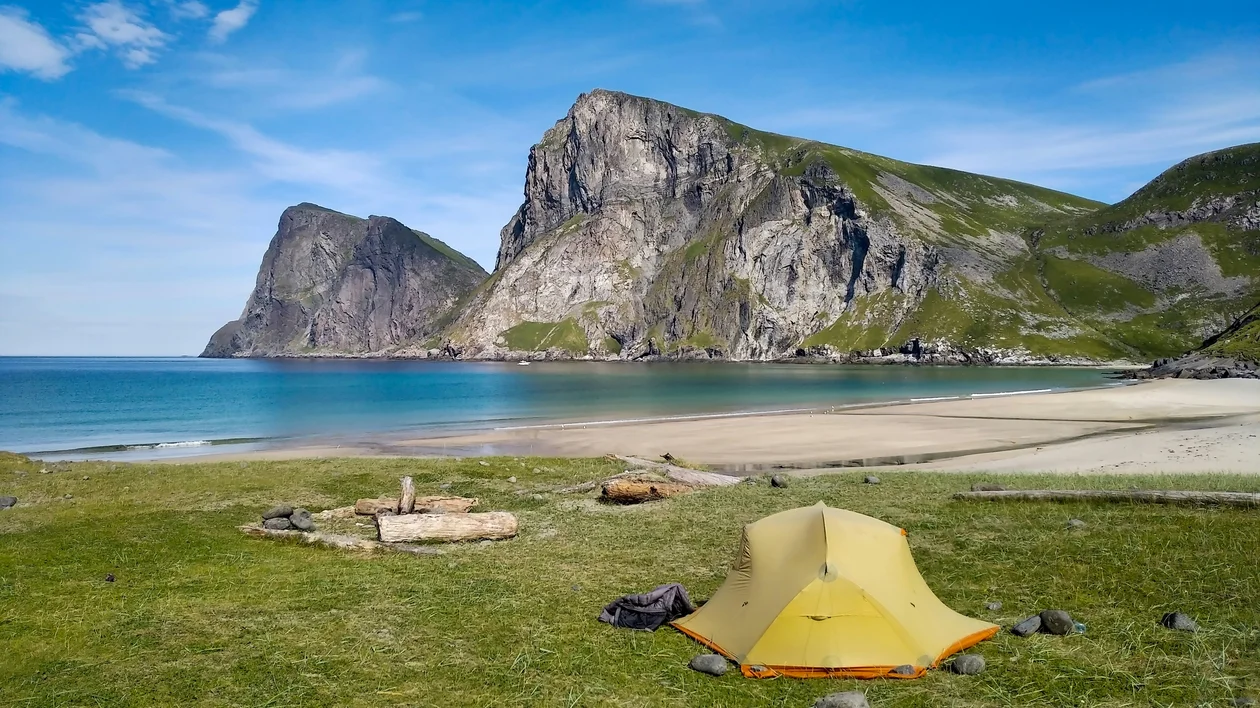 Yellow tent on grassy shore with mountains and blue sky in the background.