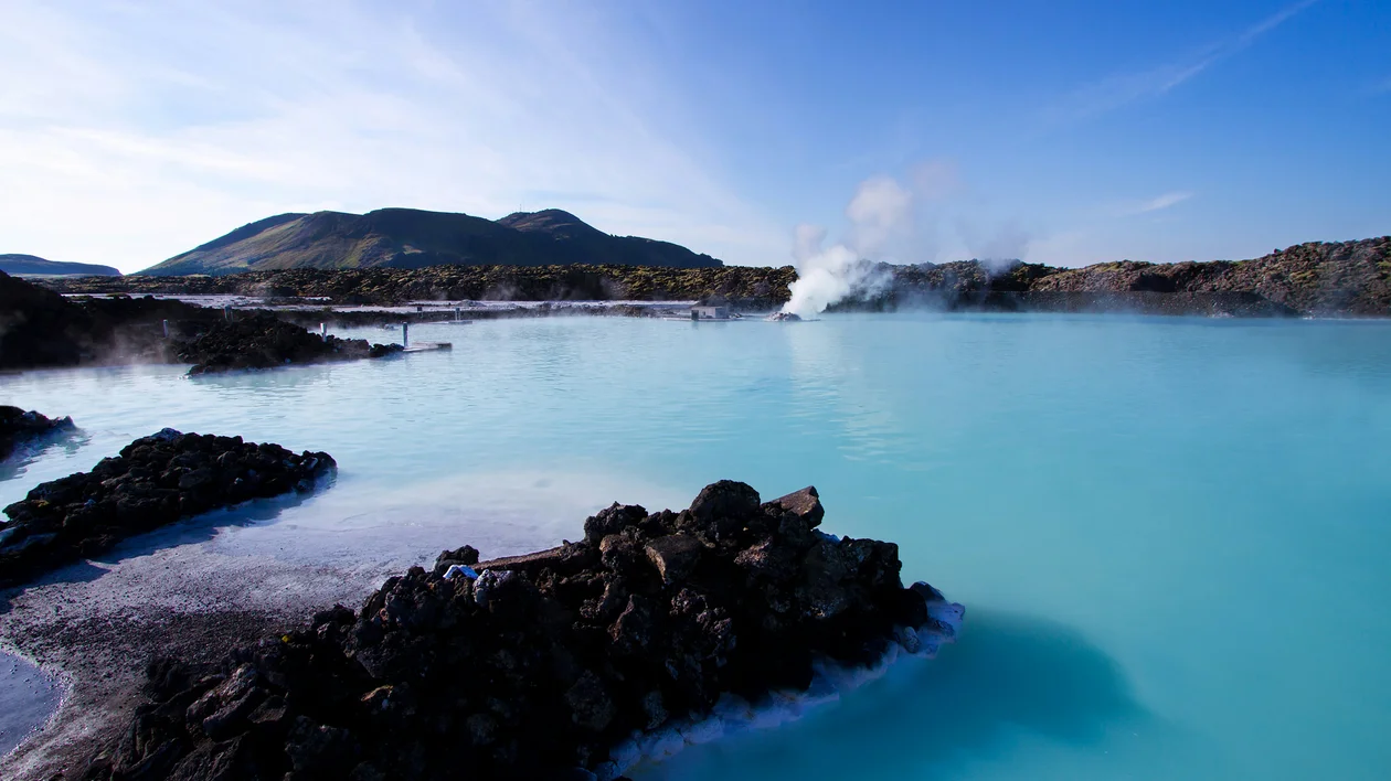 Blue geothermal lagoon with steam rising, surrounded by black rocks and distant hills.