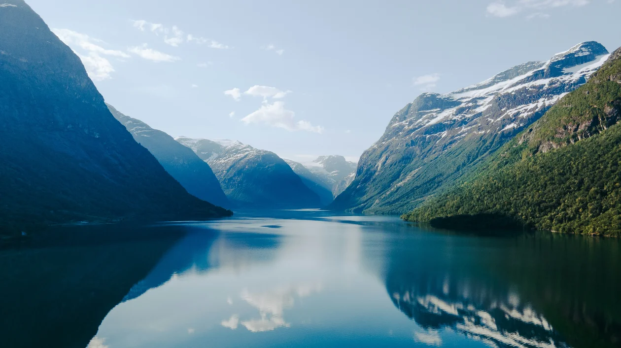 Calm lake surrounded by green mountains and snowy peaks under a clear blue sky.