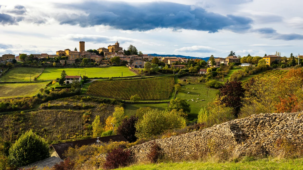 Hilltop village with stone buildings surrounded by green fields and trees under a partly cloudy sky.