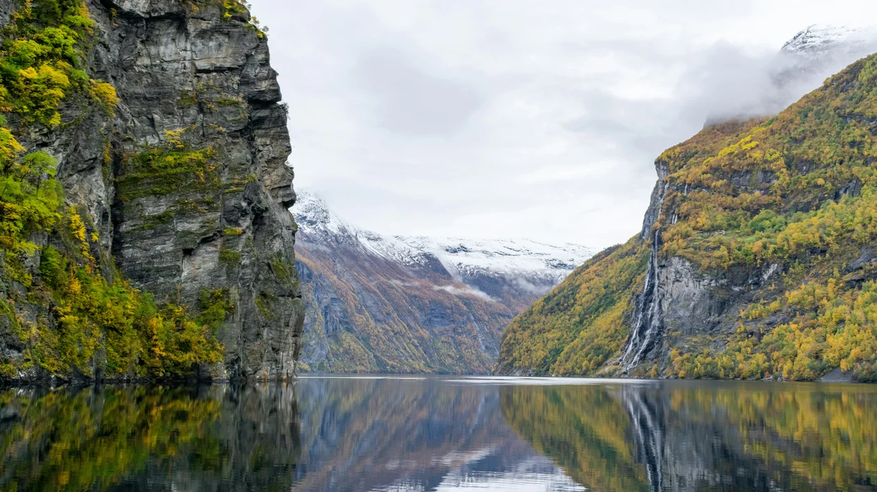 Steep cliffs and autumn trees reflected in calm fjord water under a cloudy sky.