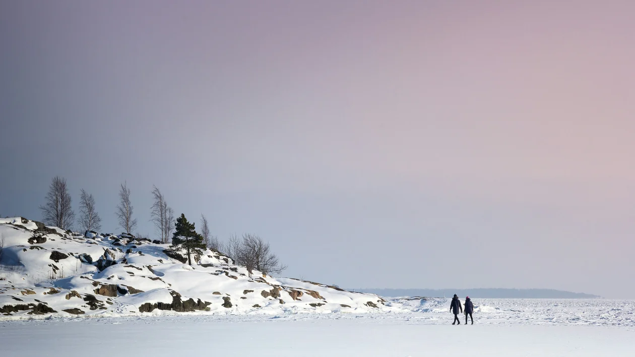 Two people walk on a snowy landscape near a treed hill under a pink and blue sky.