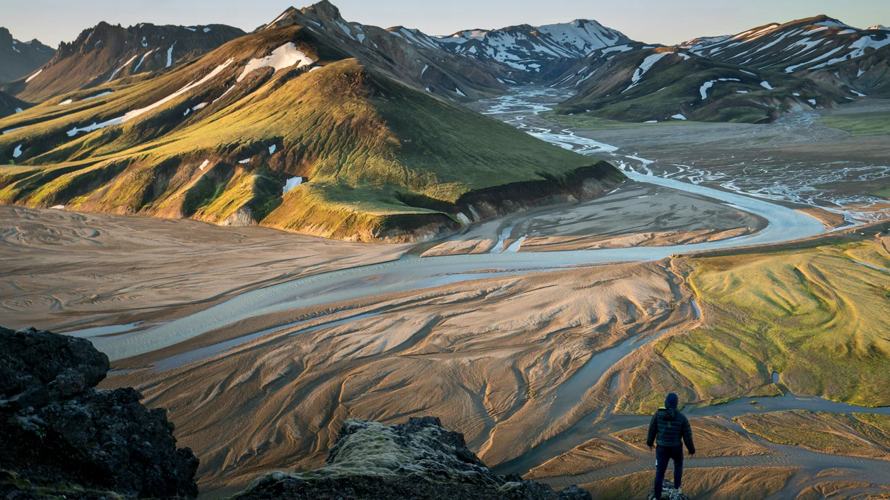 Person standing on a cliff overlooking a winding river and rugged green mountains.