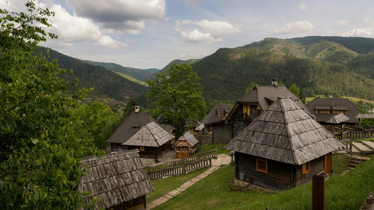 Wooden cottages with thatched roofs nestled in green mountain landscape under a partly cloudy sky.