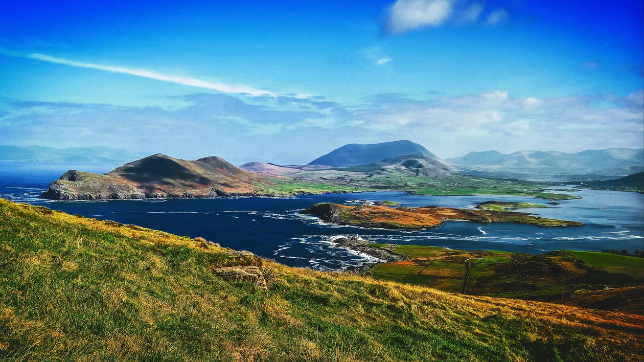 Rolling green hills overlooking a coastal village and mountains under a bright blue sky.