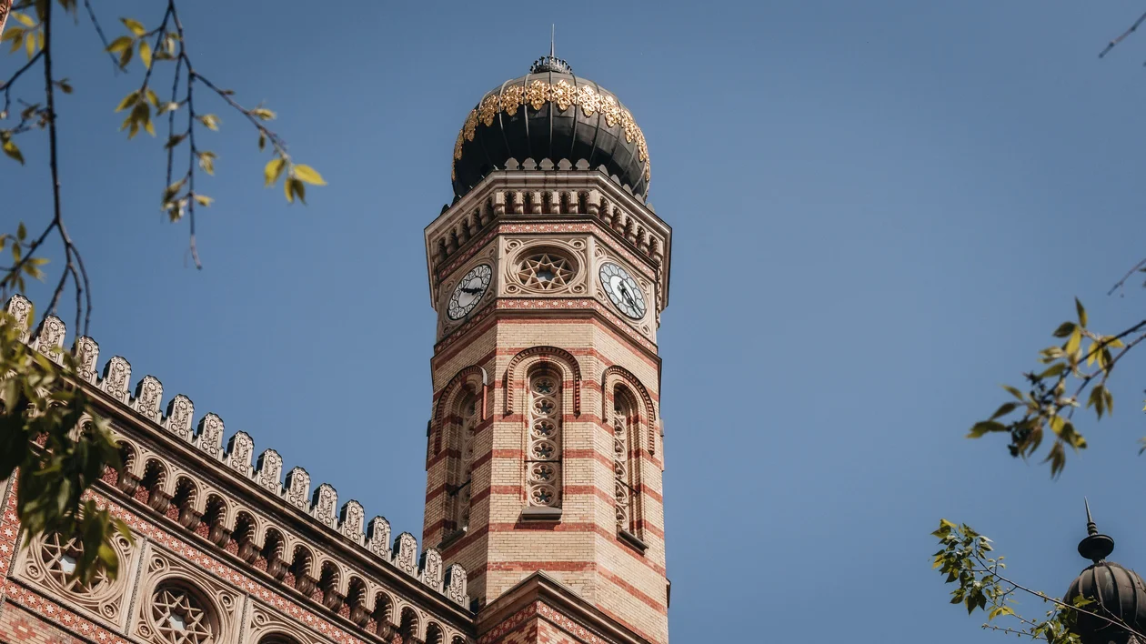 Ornate clock tower with striped brickwork against a clear blue sky, framed by tree branches.