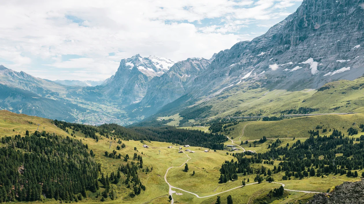 Green valley with winding trail, scattered trees, and mountains under a partly cloudy sky.