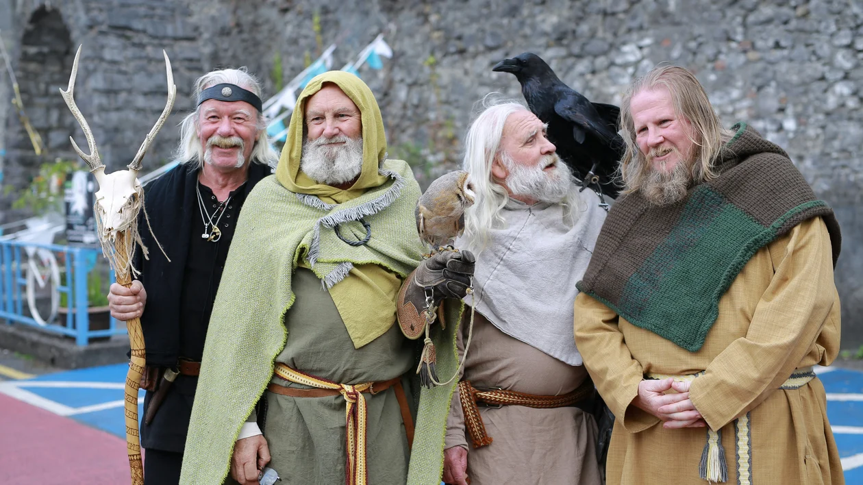 Four men in medieval costumes stand together, one with a black bird on his shoulder.
