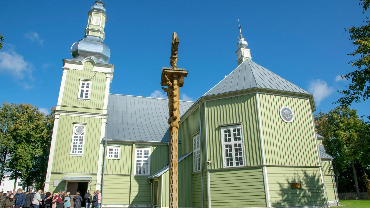 Green wooden church with a silver roof, people gathered outside under a clear blue sky.