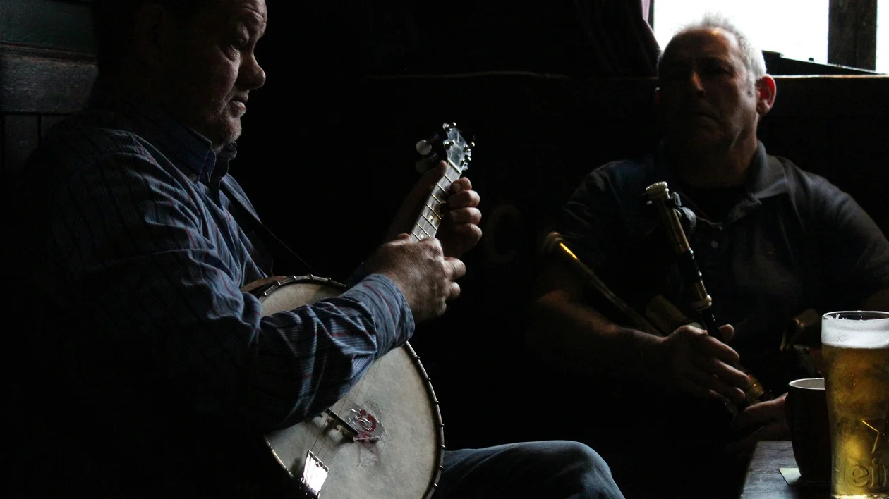 Two men play banjo and bagpipes in a dimly lit pub with a pint of beer on the table.