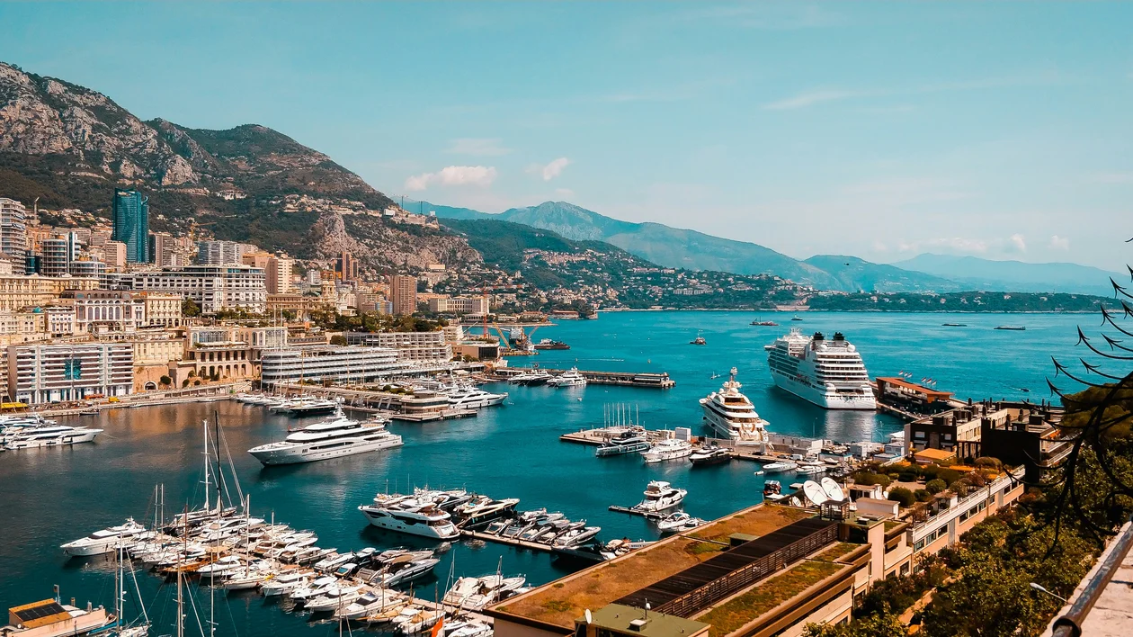 Yachts and cruise ships docked in a harbor surrounded by mountains and city buildings.