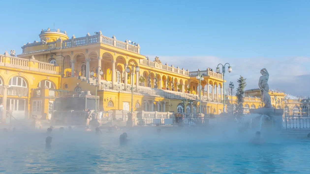 People swim in a steaming outdoor pool beside a grand yellow bathhouse under a clear sky.