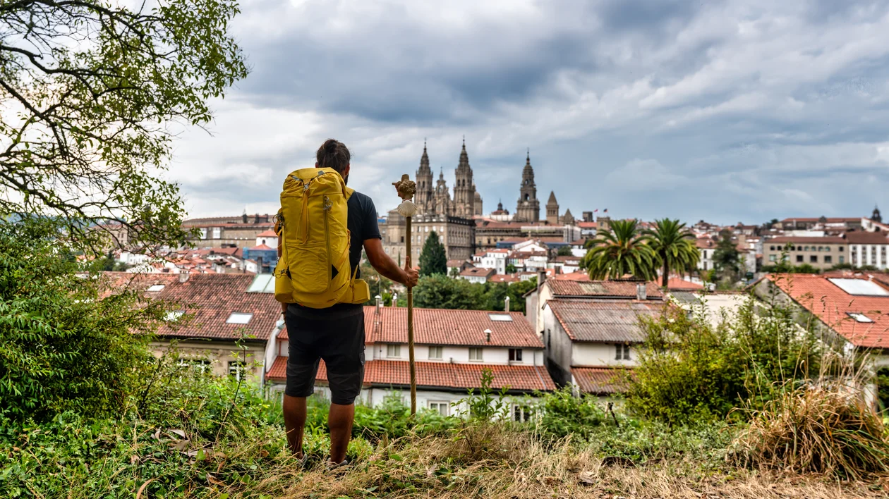Hiker with yellow backpack overlooking a city with church spires under cloudy sky.