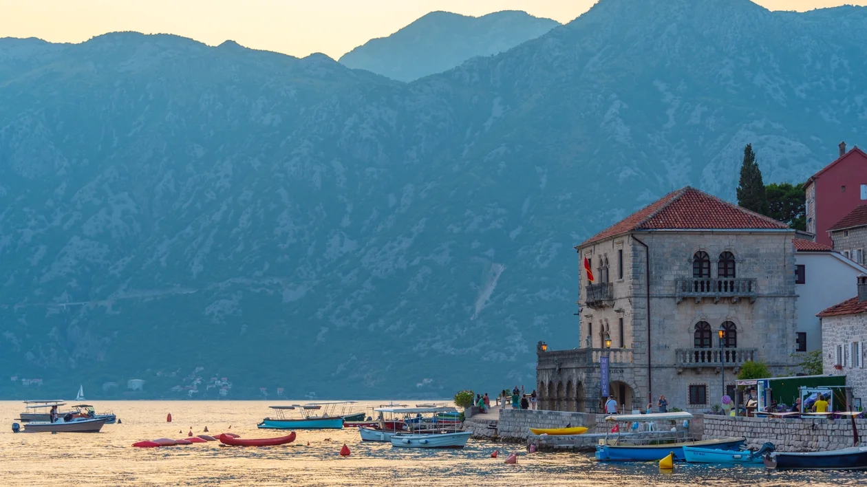 Boats on calm water near stone buildings with mountains in the background at sunset.
