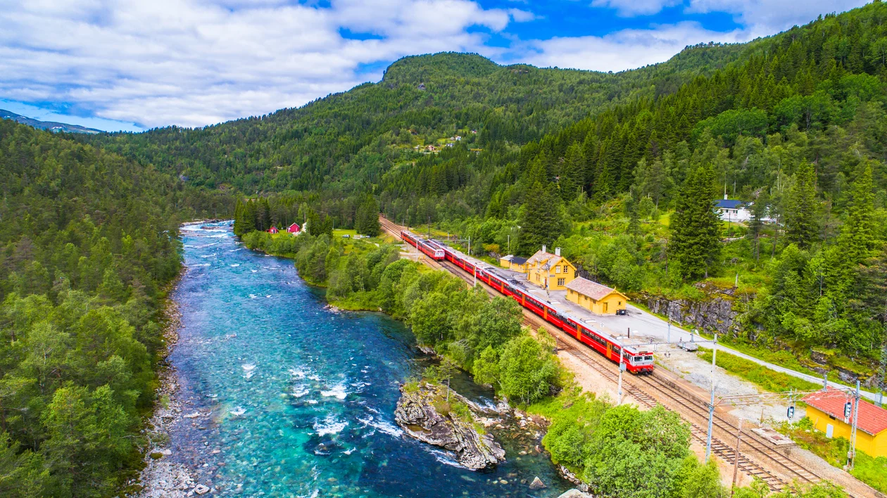 Red train passing a small yellow station by a river in a lush green mountain landscape.