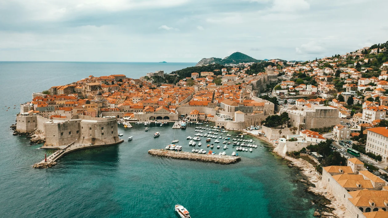 Coastal city with terracotta rooftops, a small marina, and boats in turquoise water.