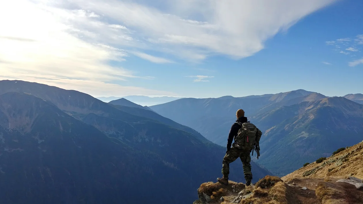 Hiker standing on a mountain ridge overlooking distant blue peaks under a bright sky.