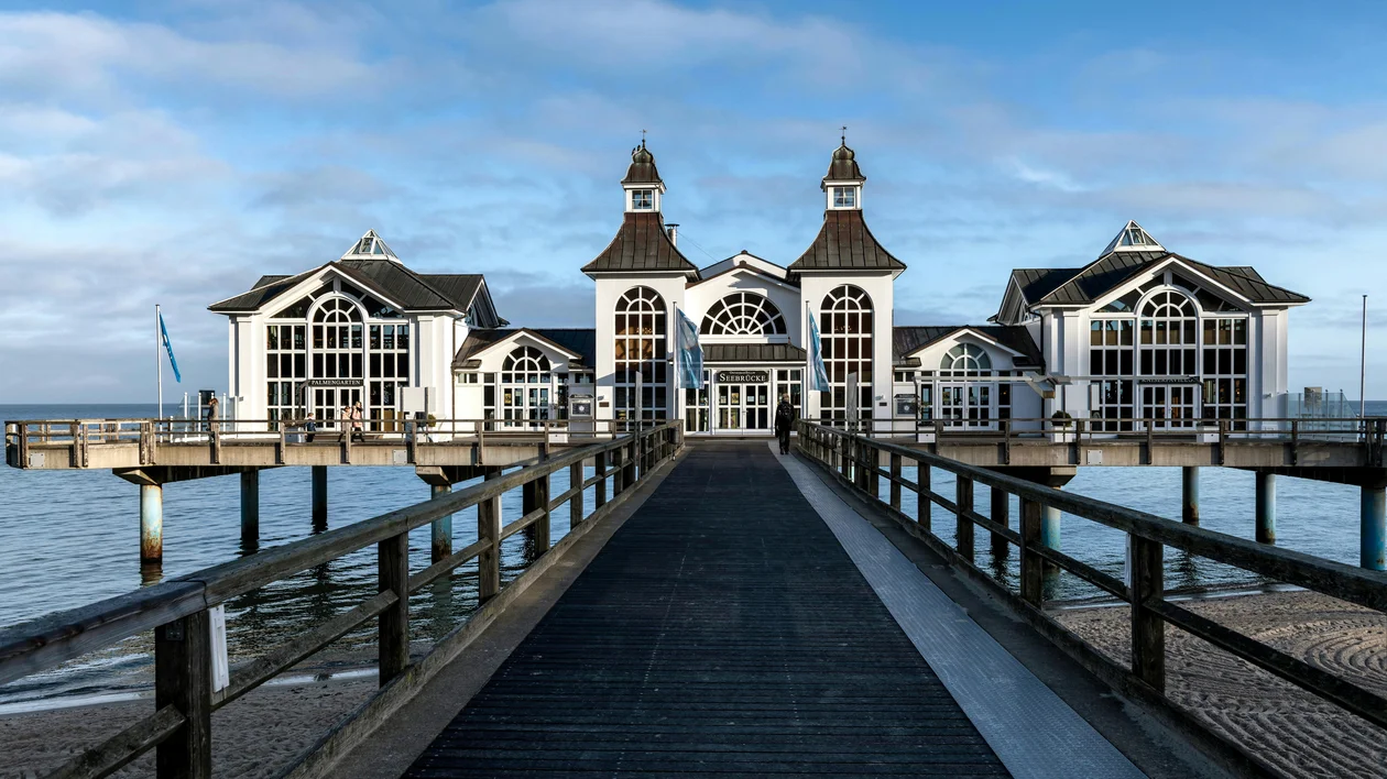 Pier with a large building over the ocean under a blue sky with scattered clouds.