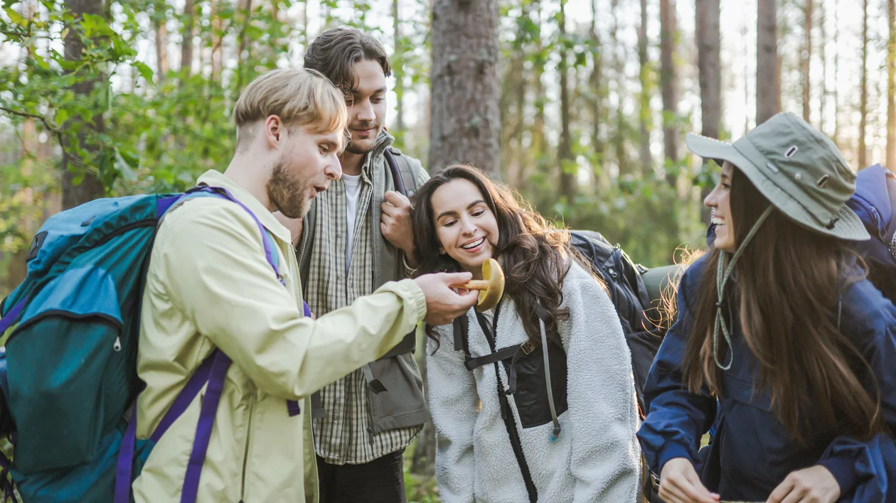 Four friends in hiking gear laughing and sharing food in a sunlit forest.