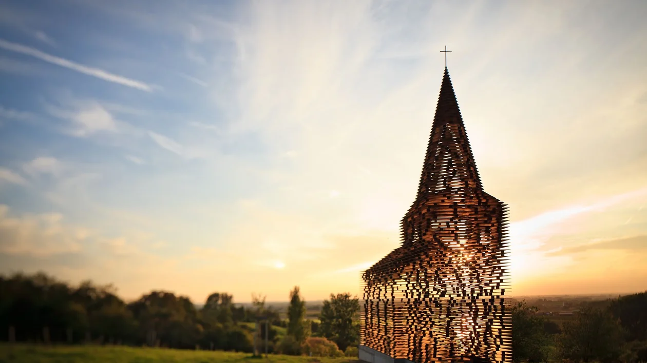 Transparent church sculpture in a field at sunset with trees in the background.