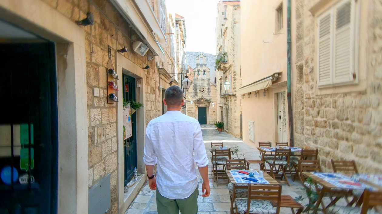 Man walking through a narrow cobblestone street lined with empty outdoor café tables.