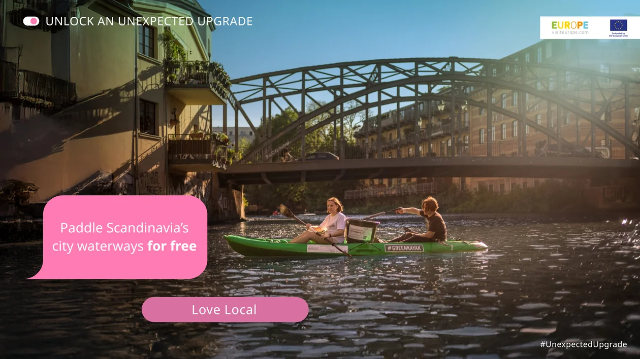 Two people kayaking on a sunlit city river with a bridge in the background.