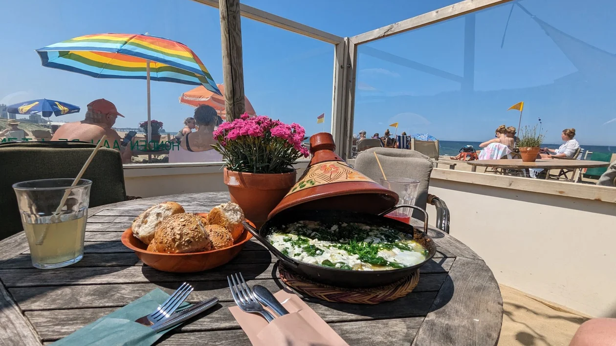 Outdoor table with bread, a tajine dish, flowers, and lemonade by a sunny beach scene.