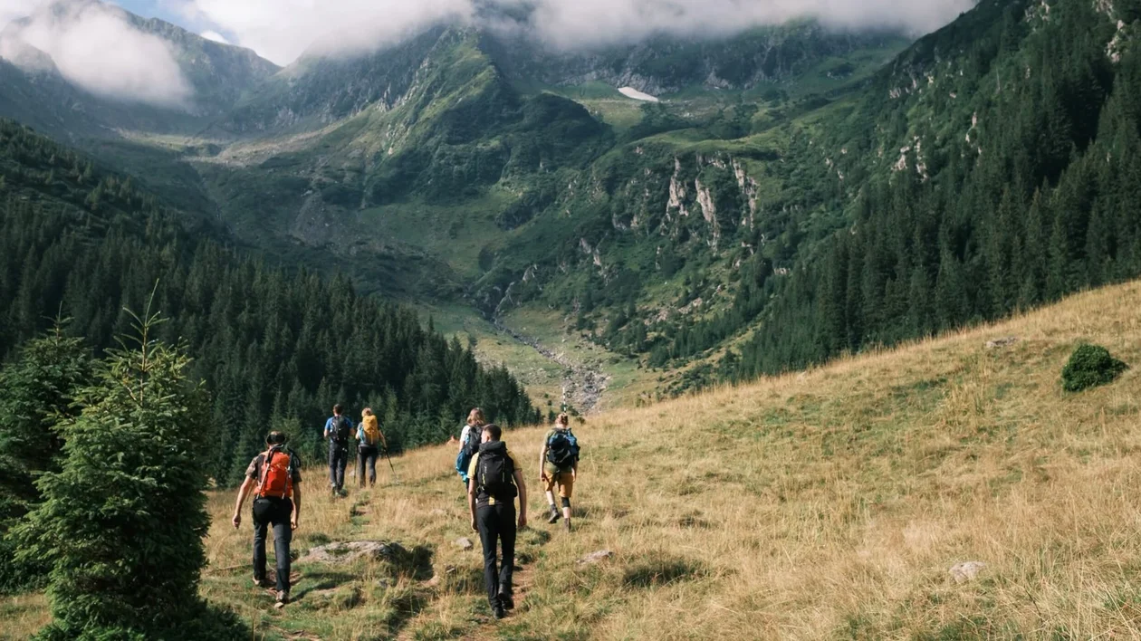 Hikers walking down a grassy mountain trail surrounded by forested hills and clouds.