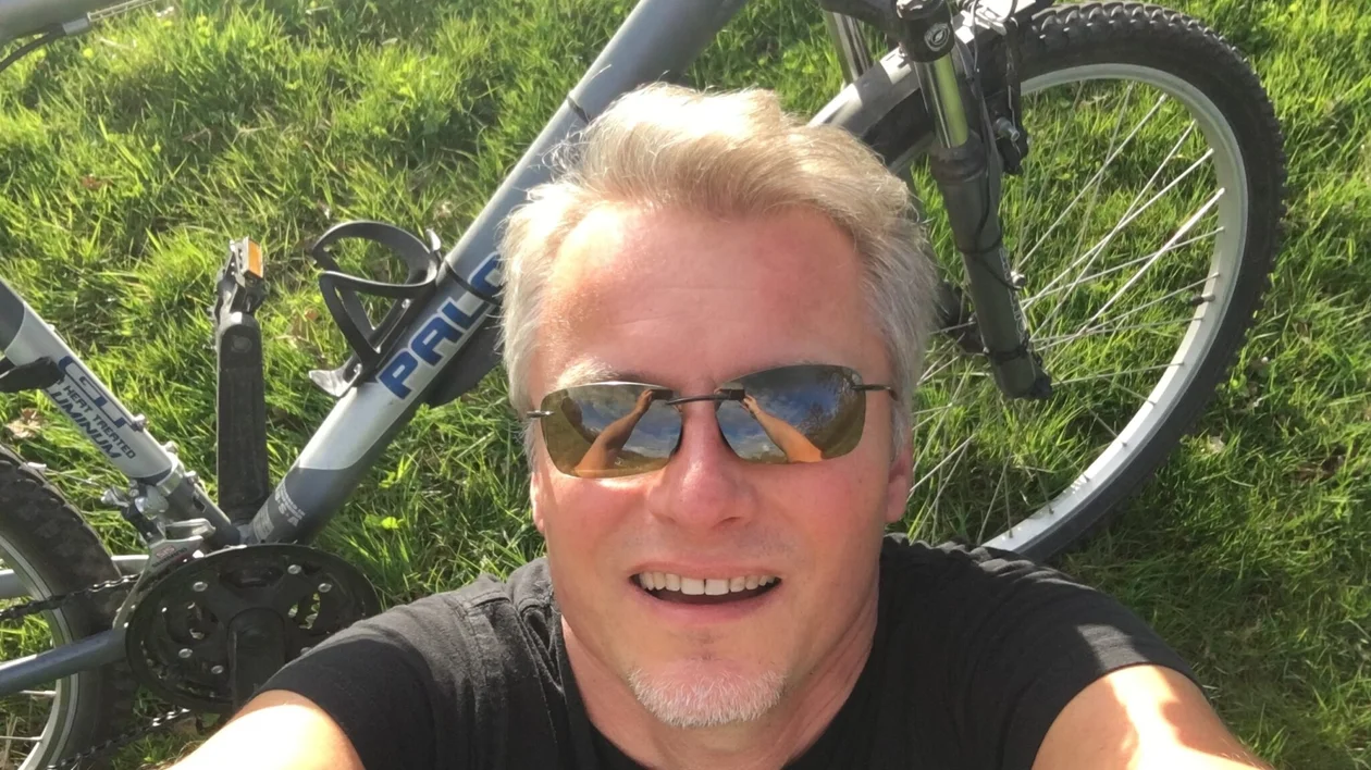 Man in sunglasses smiling outdoors next to a bicycle on green grass.