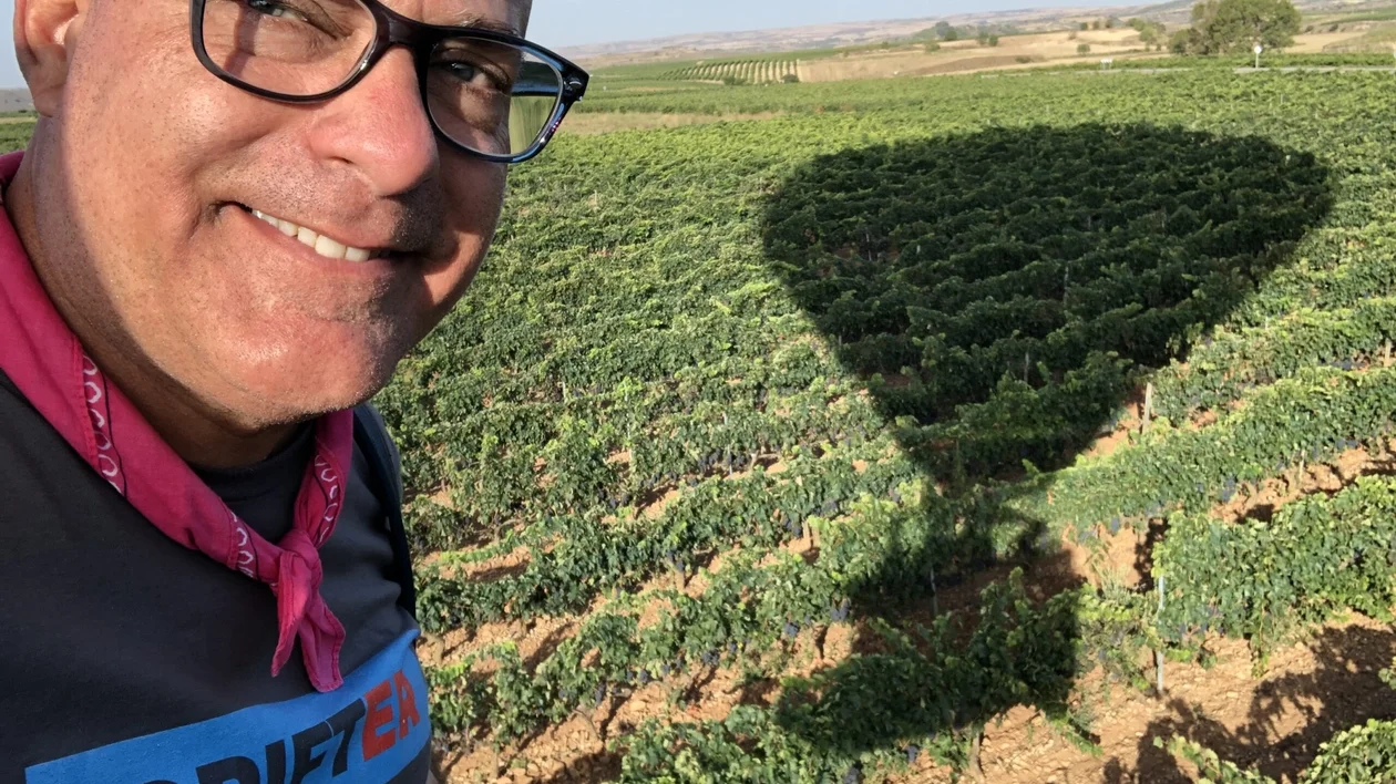 Smiling man with glasses in a field, hot air balloon shadow on greenery.