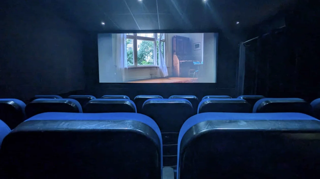 Empty movie theater with blue seats facing a screen showing a brightly lit room.