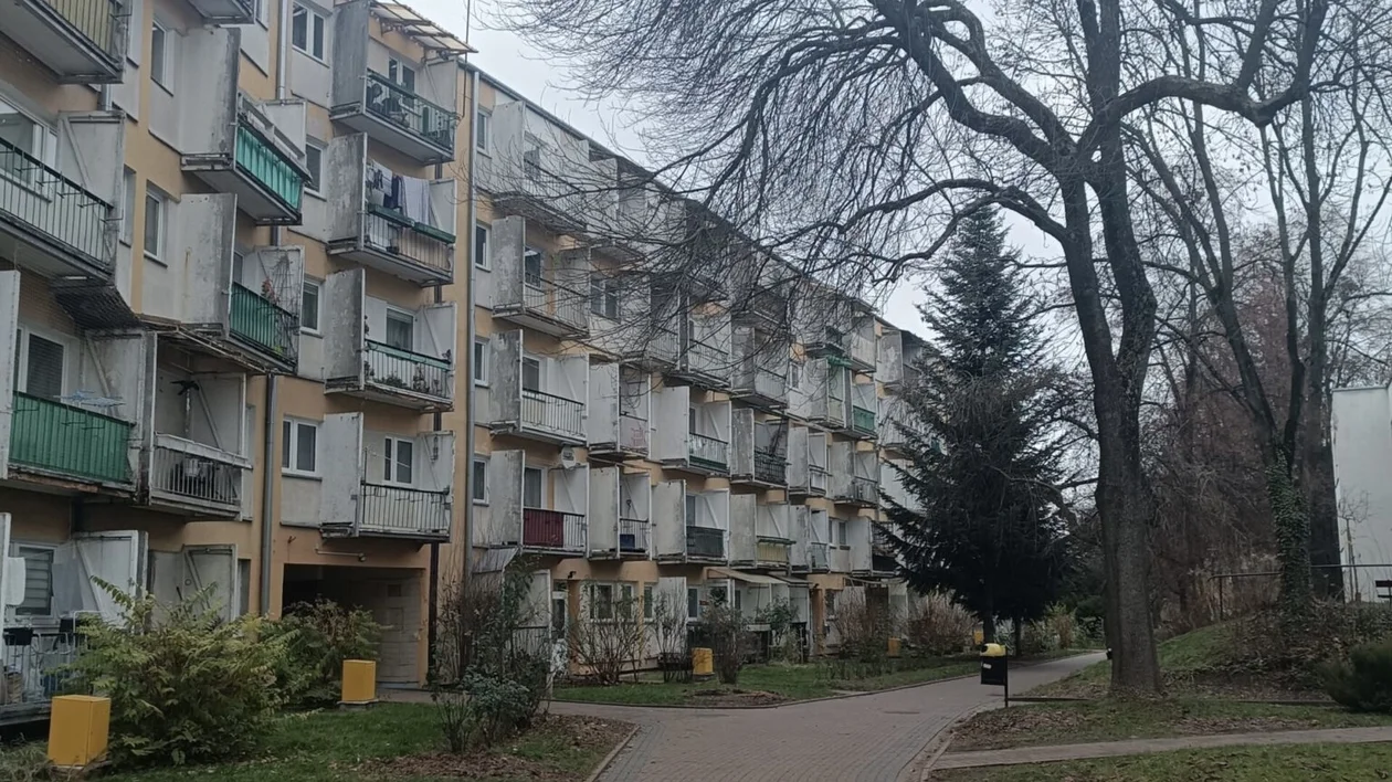 Apartment building with balconies next to a pathway and leafless trees on a cloudy day.