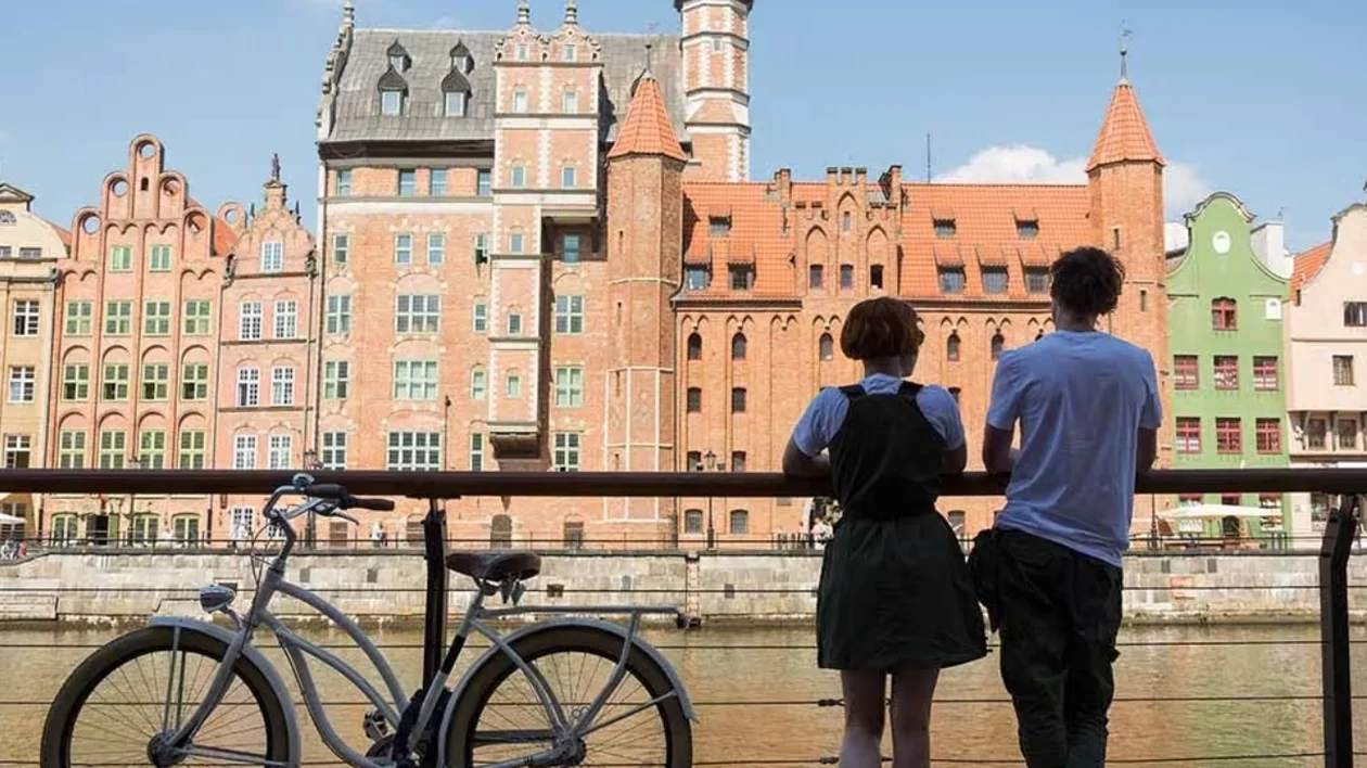 Two people and a bicycle by a river, colorful historic buildings in the background.
