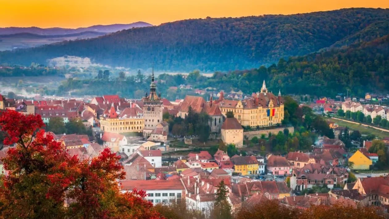 Colorful town with historic castle, autumn trees, and forested hills at sunset.