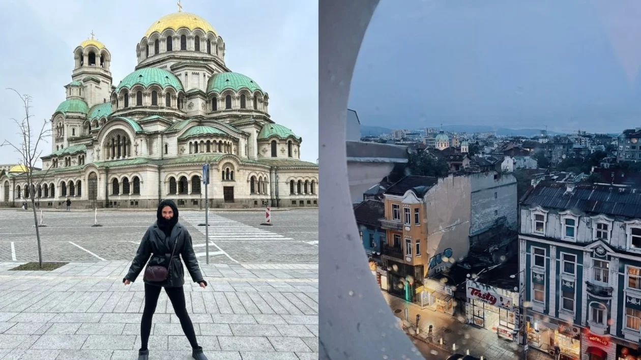 Person in black standing in front of a domed cathedral; cityscape view through a round window.