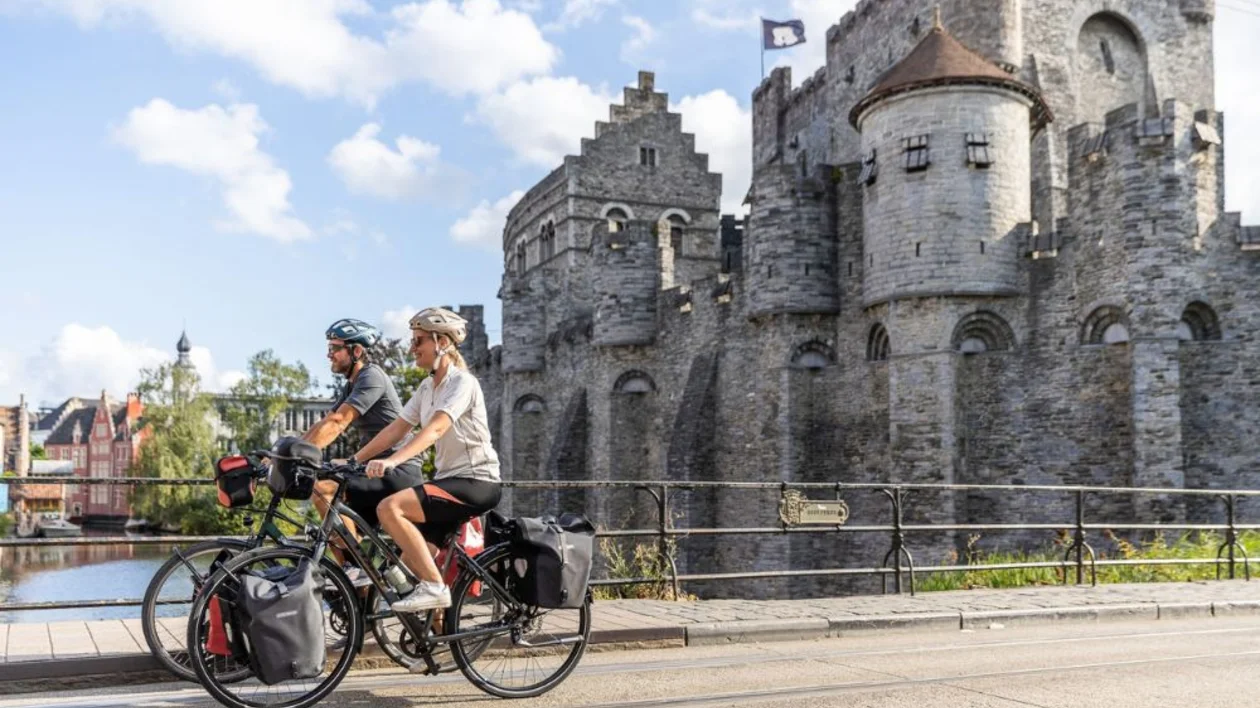 Two cyclists ride past a medieval stone castle on a sunny day.