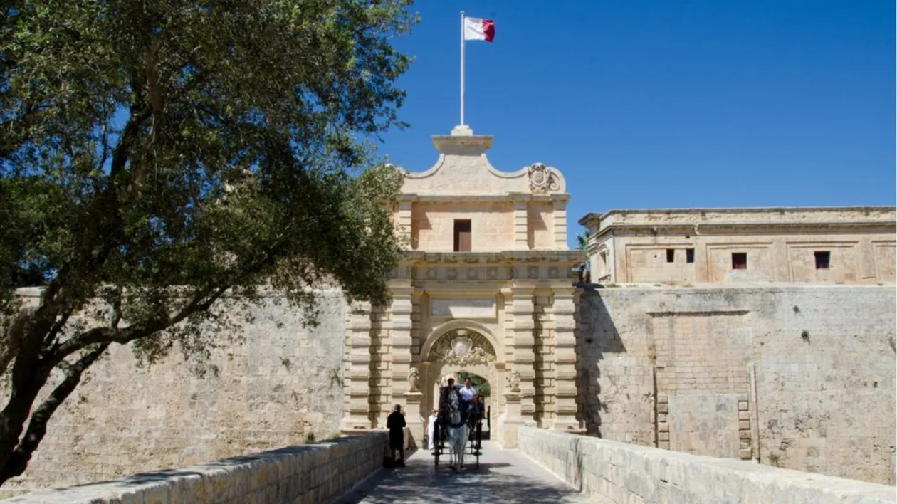 Stone fortress entrance with arched gate, Maltese flag, and people walking on bridge.
