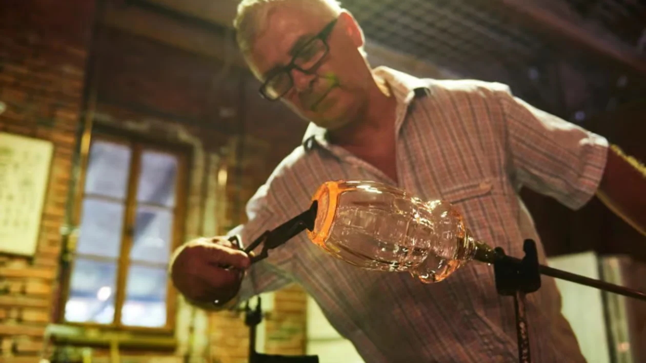 Man shaping molten glass with tools in a workshop.