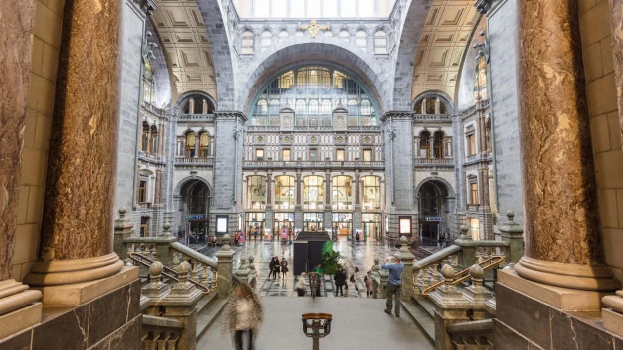 Grand hall of a train station with marble columns and arched windows, people walking.