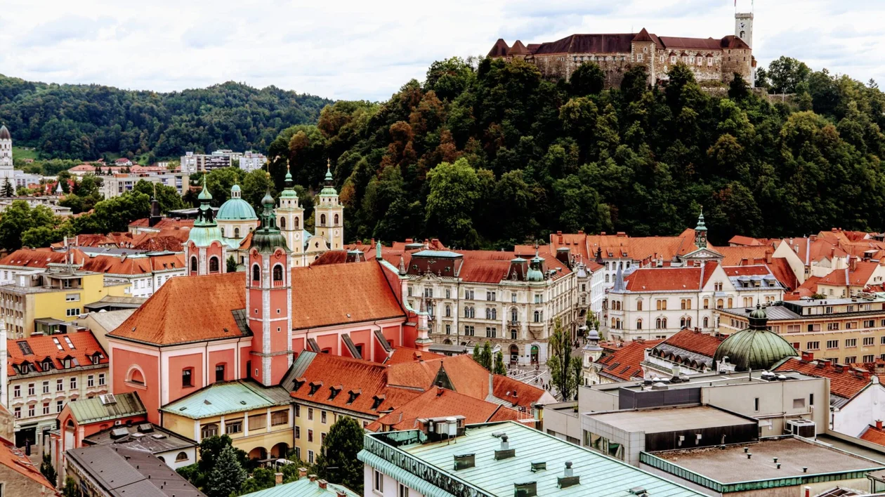 Red-roofed city buildings with a church in front and a hilltop castle surrounded by trees.