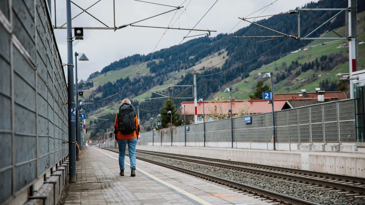 Person with backpack standing on a train platform near mountains on a cloudy day.