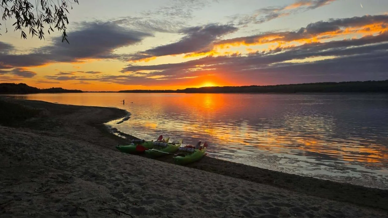 Colorful sunset over a calm lake with kayaks on a sandy shore.
