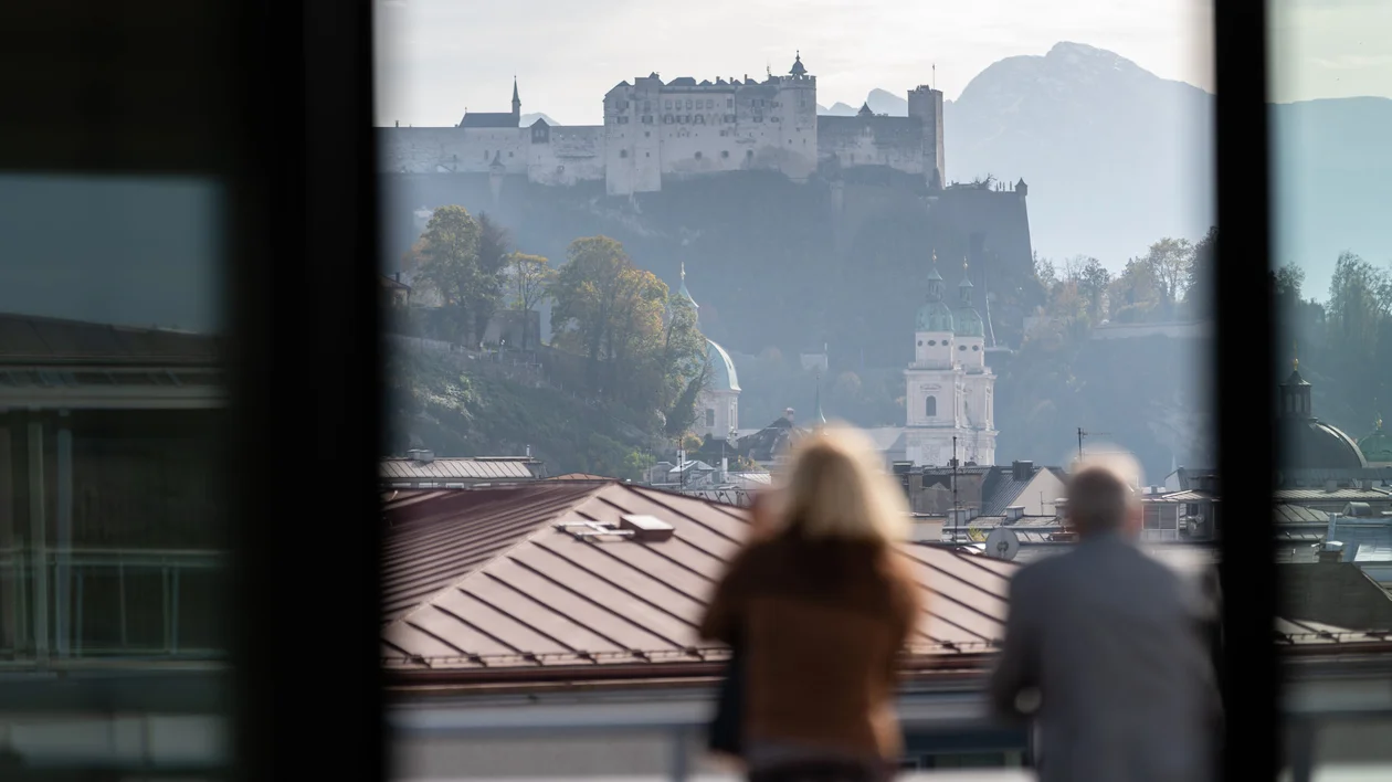 Two people on a balcony overlooking a castle and distant mountains.