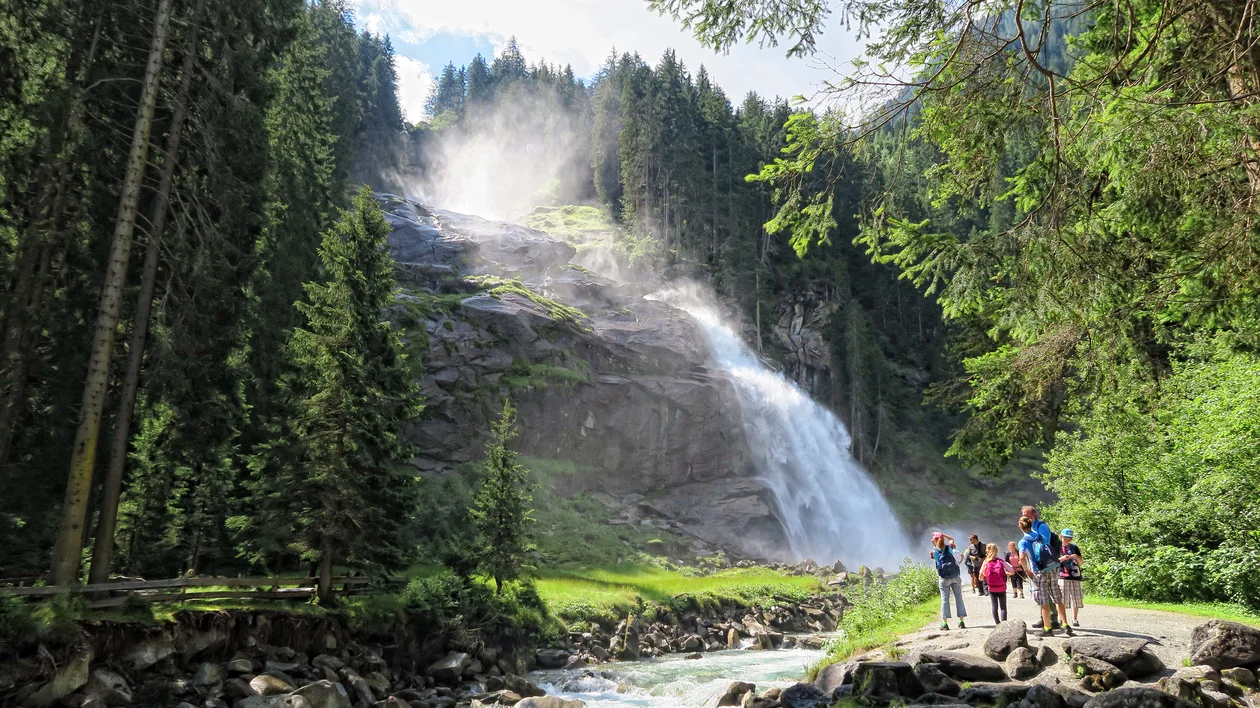 People hiking near a waterfall surrounded by tall trees and rocky stream.