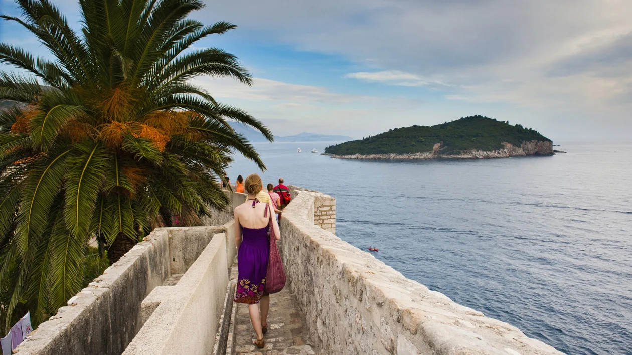 Woman walking along a stone pathway by the sea with a palm tree and island in the distance.