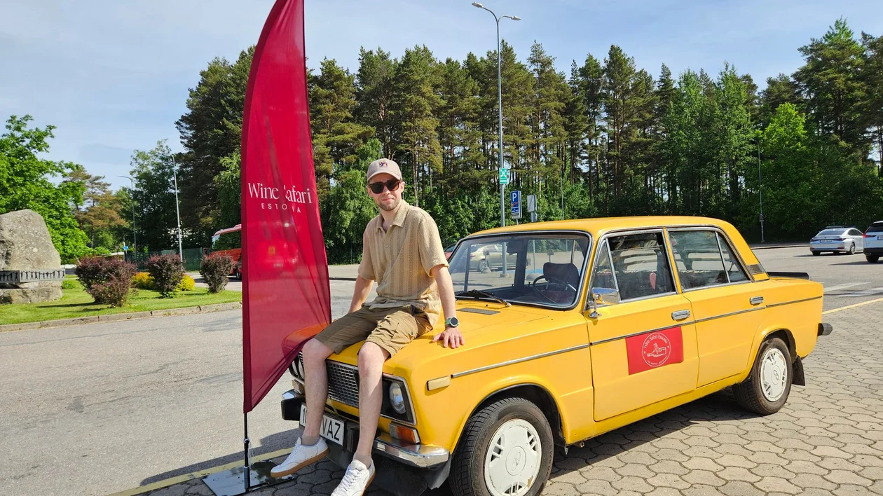 Man in sunglasses sits on yellow vintage car near a red flag on a sunny day.