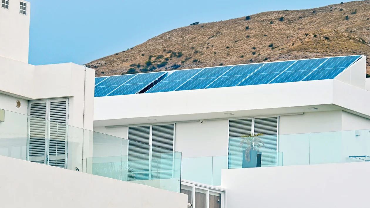 Modern white building with solar panels on the roof, glass balconies, and a hill in the background.