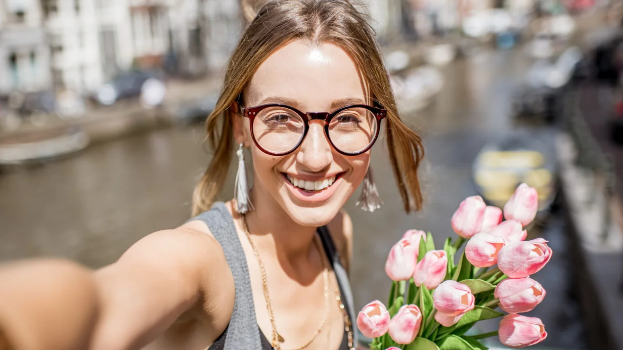 Smiling woman with glasses holding pink tulips by a canal on a sunny day.