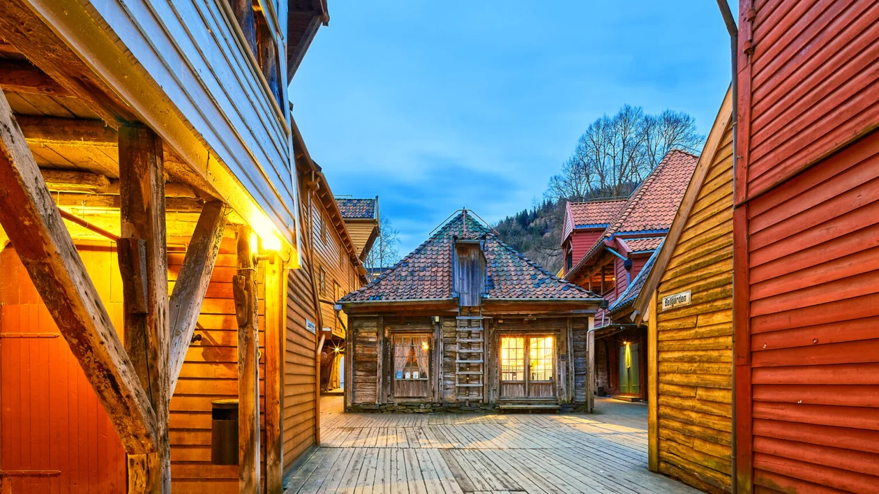 Narrow wooden alley with colorful timber buildings at dusk, warm lights glowing inside.